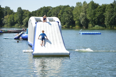 Junge rutscht von einer aufblasbaren Rutsche ins Wasser, während ein Mädchen oben sitzt und andere Badegäste im Hintergrund sind.