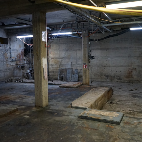 Leere, halbdunkle Betonhalle mit Stahlpfeilern, Kabeln und unsauberen Böden.