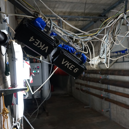 Zwei schwarze Steuerkästen mit Beschriftung hängen von Kabeln in einem grauen Kellerraum mit Rohren.