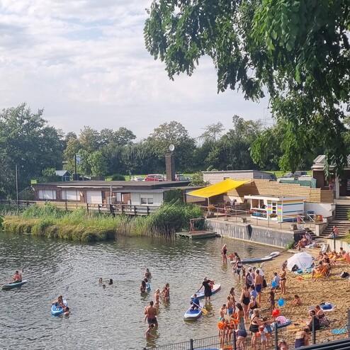 Blick auf ein Gewässer mit Badegästen, Booten und einem Sandstrand vor einem bunten Restaurant mit Sonnenschirmen.