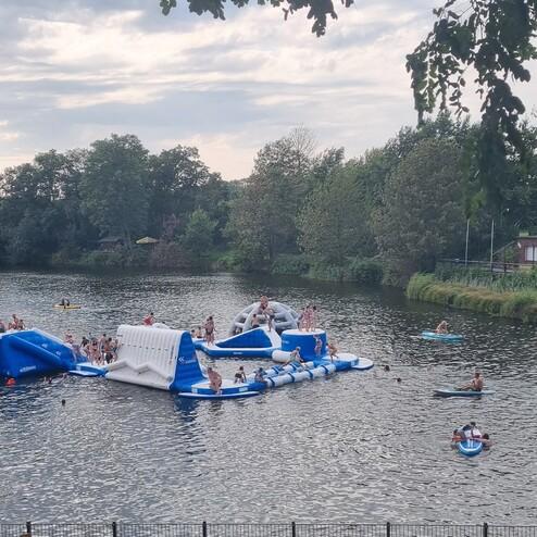 Menschen schwimmen und spielen auf einem Wasser-Spielplatz in einem See, umgeben von Bäumen und einem ruhigen Ufer.