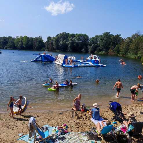 Strandtag mit Menschen am Wasser, Spielgeräten, Surfbrettern und Liegen auf sandigem Ufer unter blauem Himmel.