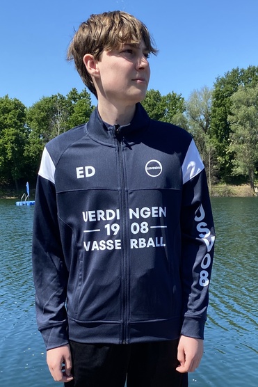 Junger Mann steht auf einem Holzsteg am Wasser, trägt einen Sportanzug mit dem Aufdruck "VASSERBALL".