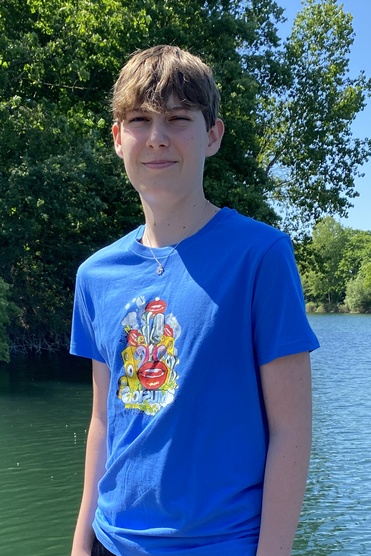 Ein Junge steht auf einem Holzsteg am Wasser, trägt ein blaues T-Shirt und schwarze Shorts, umgeben von Bäumen.