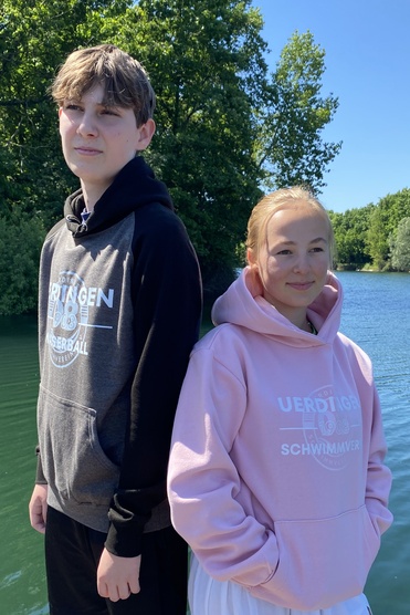 Zwei Jugendliche stehen auf einem Holzsteg am Wasser, umgeben von grünen Bäumen und klarem Himmel.