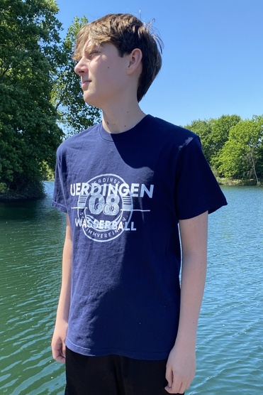 Junger Mann steht auf einem Steg am Wasser, trägt ein dunkelblaues T-Shirt und kurze schwarze Hosen, im Hintergrund Bäume.