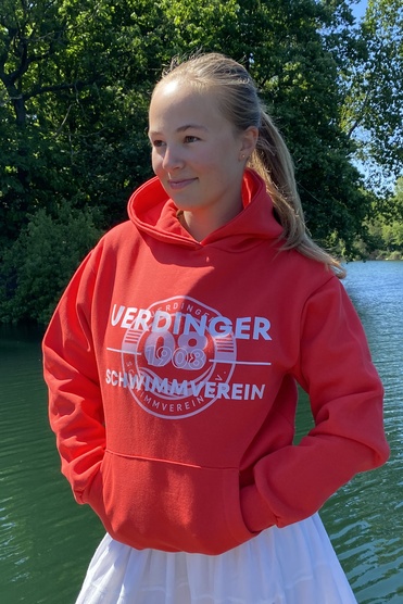 Junge Frau in rotem Hoodie und weißem Rock steht am Ufer eines Gewässers, umgeben von grünen Bäumen.