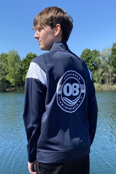 Junge in Sportbekleidung mit Logo steht am Ufer eines Gewässers unter blauem Himmel.