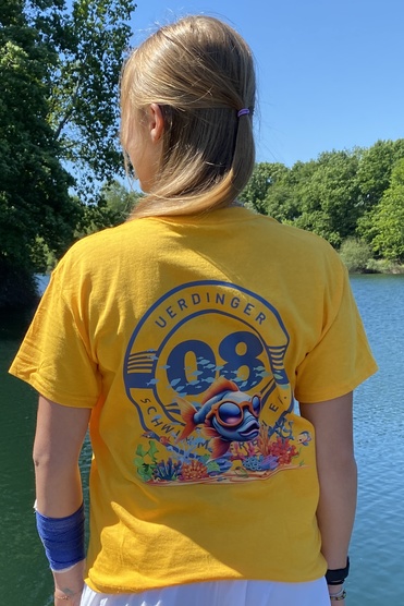 Mädchen in gelbem T-Shirt mit Motiv steht am Wasser, umgeben von Bäumen, blickt auf die ruhige Oberfläche des Sees.