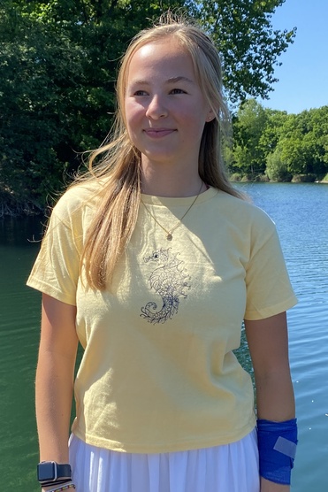 Junge Frau mit langen Haaren in gelbem T-Shirt und weißem Rock steht am Wasserrand eines ruhigen, grün umgebenen Sees.