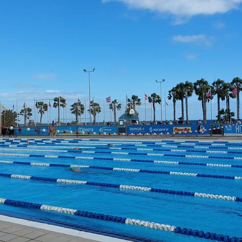 Schwimmbecken mit blauen und weißen Bahnen, umgeben von Palm trees und Flaggen vor einem Bergpanorama.