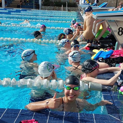 Gruppenszenario im Schwimmbecken mit Jugendlichen in Schwimmkappen, einige halten Schwimmhilfen, eine Person zeigt Daumen hoch.