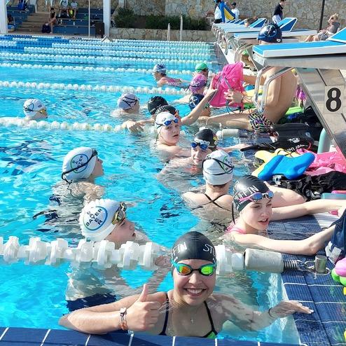 Gruppe von Schwimmern im Wasser eines Schwimmbeckens, einige lächelnd, mit Badekappen und Schwimmbrillen.