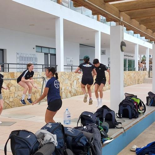 Sechs Mädchen springen auf einer Sportfläche, während Taschen und Wasserflaschen im Hintergrund liegen.