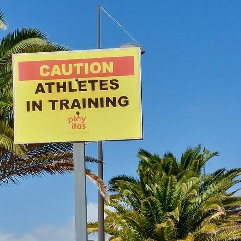 Warnschild "Caution Athletes in Training" vor Palmen und blauem Himmel.