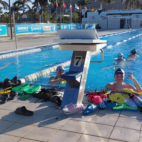 Schwimmerinnen und Schwimmer entspannen am Beckenrand eines Schwimmbeckens, umgeben von Trainingsutensilien und Sonnenlicht.