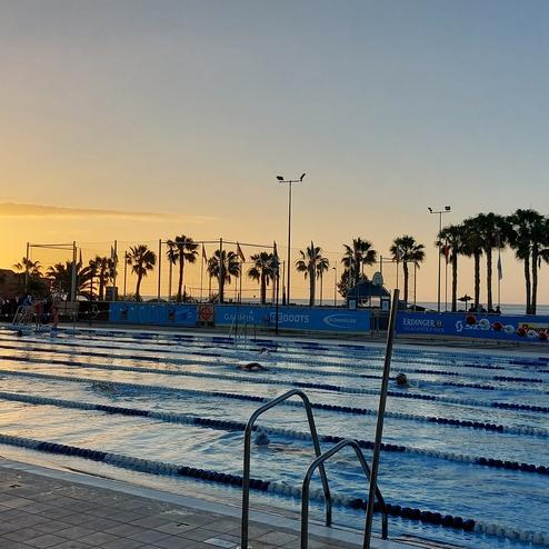 Schwimmbad mit mehreren Bahnen und Palmen, im Hintergrund ein Sonnenuntergang über den Bergen.
