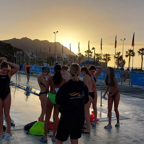 Gruppenszenen von Schwimmern bei Sonnenuntergang am Pool mit Palmen und Wettkampfbanden im Hintergrund.