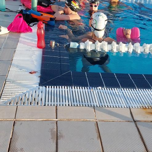 Gruppen von Schwimmern mit Platten und Wasserflaschen stehen am Beckenrand eines Schwimmbades während des Trainings.