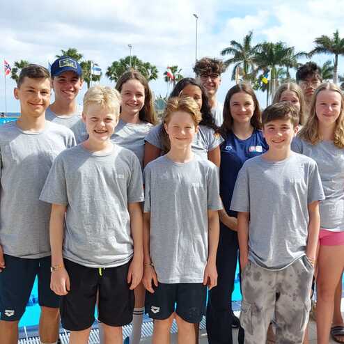Gruppe von Schwimmern in grauen T-Shirts vor einem Schwimmbecken, umgeben von Palmen und Flaggen.