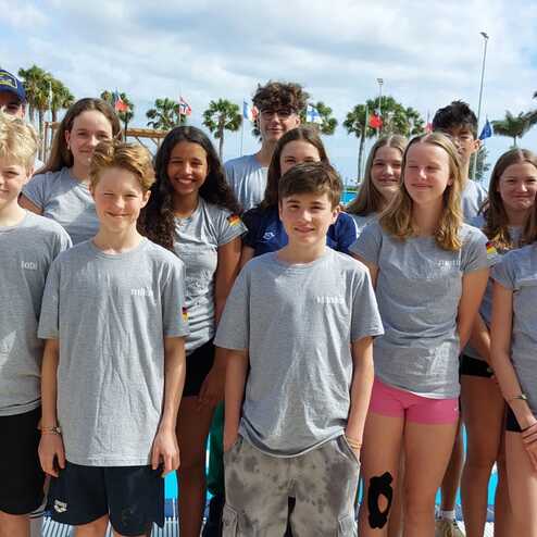 Gruppe junger Schwimmer in grauen T-Shirts vor einem Schwimmbecken, umgeben von Palmen und Flaggen.