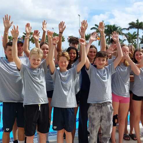 Gruppe von Schwimmern in grauen T-Shirts hebt die Hände vor einem Schwimmbecken mit Palmen im Hintergrund.