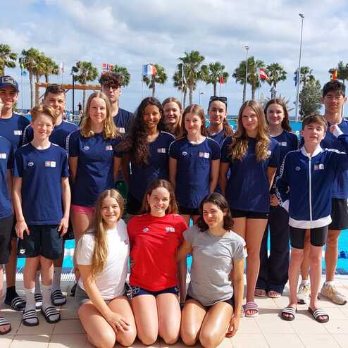 Gruppe von Schwimmern in Sportkleidung, posierend vor einem olympischen Schwimmbecken mit Palmen im Hintergrund.