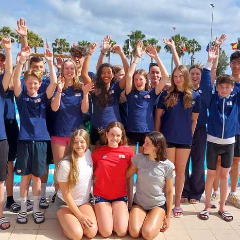 Gruppe von Schwimmern posiert lächelnd vor einem Schwimmbecken unter Palmen, einige heben die Arme zur Feier.