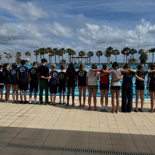 Gruppe von Schwimmern steht am Poolrand und blickt auf die Schwimmbecken, mit Palmen und Fahnen im Hintergrund.