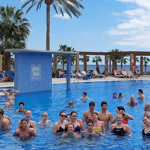 Gruppe von Menschen im Wasser eines Pools, umgeben von Palmen und Liegen, bei sonnigem Wetter.