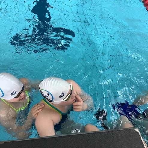 Zwei Kinder mit Schwimmmützen schwimmen zusammen im Wasser eines Schwimmbads. Ein Beckenrand ist im Vordergrund sichtbar.