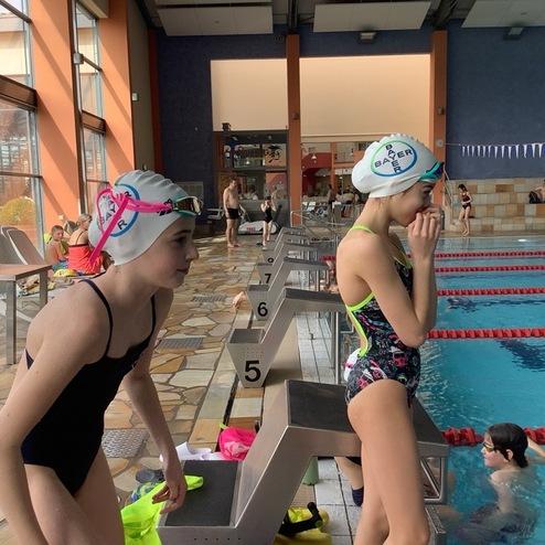 Zwei junge Schwimmerinnen warten am Beckenrand eines Schwimmbades, eine trägt einen weißen Badekapp.