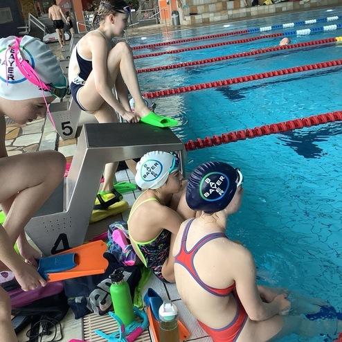 Vier Schwimmerinnen in Badeanzügen und Badekappen sitzen am Beckenrand eines Schwimmbads, bereit zum Training.