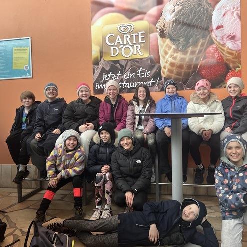 Gruppe von Kindern in Winterkleidung sitzt um einen Tisch, mit einer Eisdiele im Hintergrund.