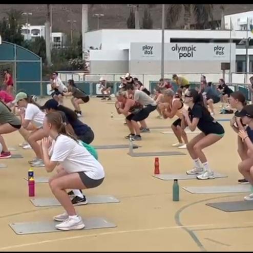 Gruppe junger Menschen in Sportkleidung macht Kniebeugen auf einer Sportfläche mit Matten und Wasserflaschen.