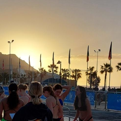 Gruppierung junger Schwimmerinnen am Beckenrand vor Sonnenuntergang, umgeben von Palmen und Nationalflaggen.