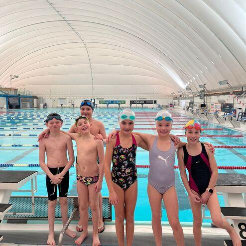 Sechs Kinder in Badekleidung und Schwimmkappen posieren am Beckenrand in einem Schwimmbad.