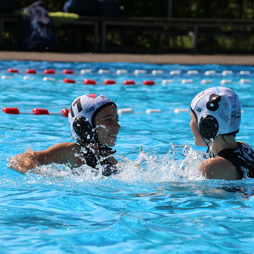Zwei Wasserballspielerinnen im Wasser l&auml;cheln sich an, spritzen Wasser auf einer hellblauen Wasseroberfl&auml;che.