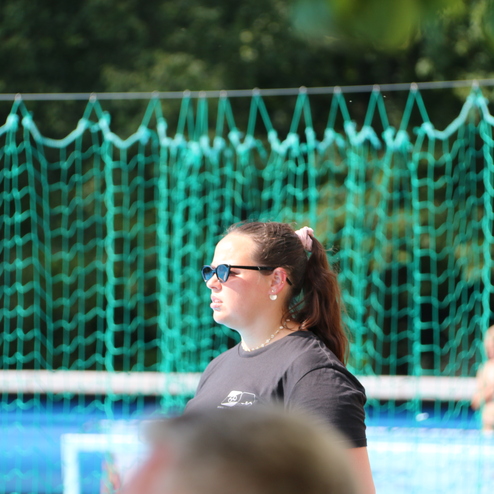 Frau mit Sonnenbrille und schwarzem T-Shirt steht vor einem gr&uuml;nen Netz in einem Schwimmbad. Im Hintergrund sind Kinder zu sehen.