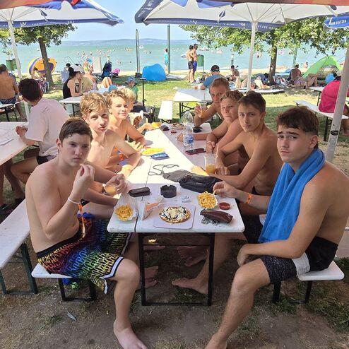Gruppe von Jungen sitzt an einem Tisch im Freien, isst Snacks und trinkt, mit Blick auf einen Badesee im Hintergrund.