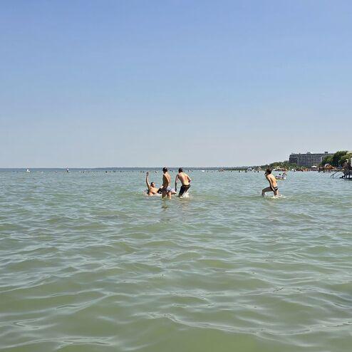 Gruppe von Menschen beim Spielen im Wasser eines Sees an einem sonnigen Tag, im Hintergrund ein Riesenrad und Strandbänke.