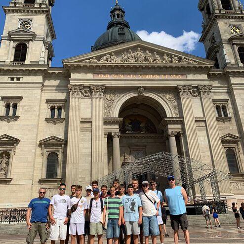 Gruppe junger Männer posiert vor der prächtigen Fassade einer Kathedrale bei strahlendem Sonnenschein.