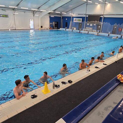 Wasserballtraining in einem Indoor-Pool mit mehreren Sportlern, die am Beckenrand stehen und sich unterhalten.