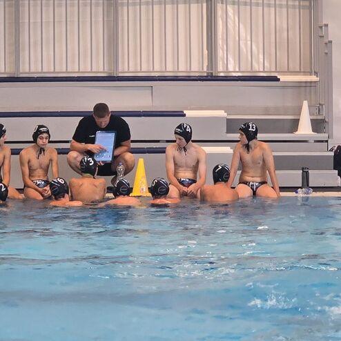 Wasserballmannschaft sitzt am Beckenrand; Trainer gibt Anweisungen, während Spieler im Wasser zuhören.
