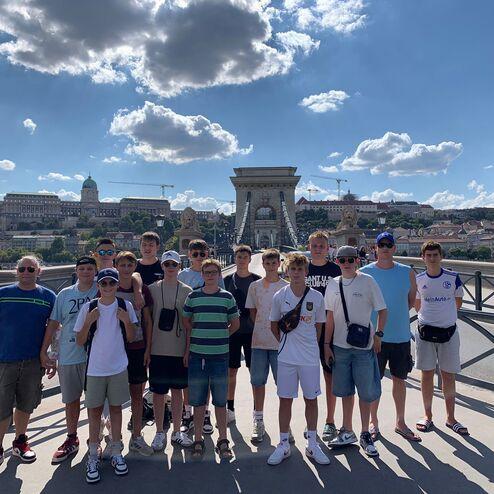 Gruppe von Jugendlichen und Erwachsenen posiert auf der Kettenbrücke in Budapest mit Blick auf historische Gebäude.