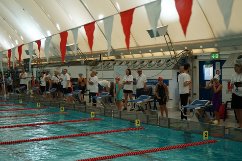 Schwimmbad mit Wettkampfschwimmern und Trainern an der Strecke, rote und wei&szlig;e Fahnen hangen von der Decke.