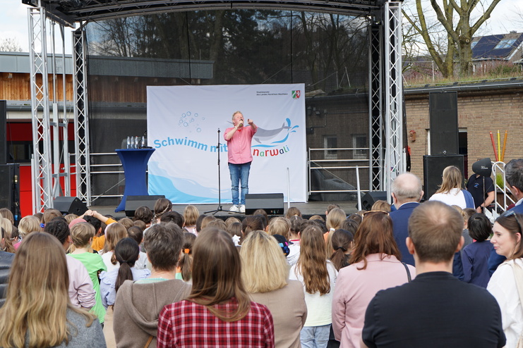 Ein Redner steht auf einer Bühne und spricht zu einer Gruppe von Kindern und Erwachsenen bei einer Veranstaltung.