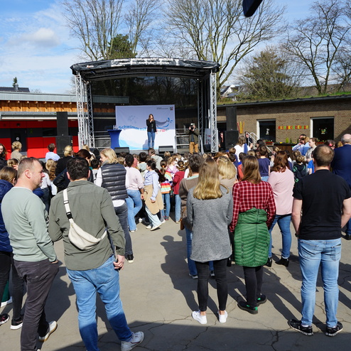 Eine Menschenmenge hört einer Rednerin auf einer Bühne in einem Außenbereich zu, unter einem blauen Himmel.