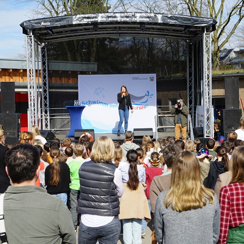Frau spricht auf einer Bühne vor einer versammelten Gruppe von Kindern und Erwachsenen bei einer Schwimmveranstaltung.