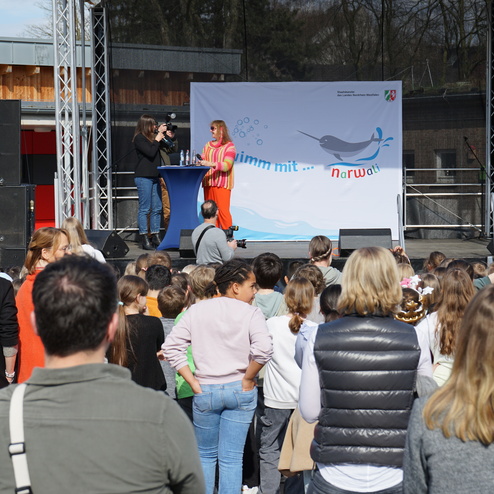 Bühne mit zwei Personen bei einer Veranstaltung vor einer Kindergruppe, Banner mit Narwal-Motiv im Hintergrund.